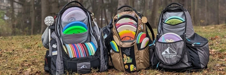 Tre Upper Park tasker der står fyldt med disc golf discs ved siden af hinanden i en skovbund